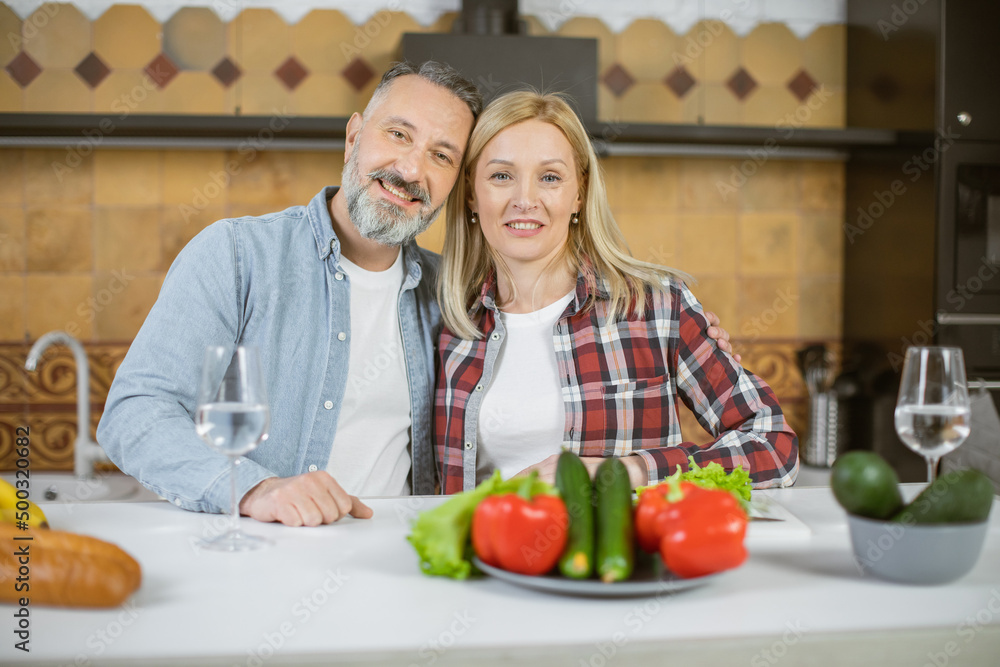 Portrait of beautiful caucasian couple in casual wear standing in embrace on bright kitchen and smiling on camera. Fresh organic food lying on table.