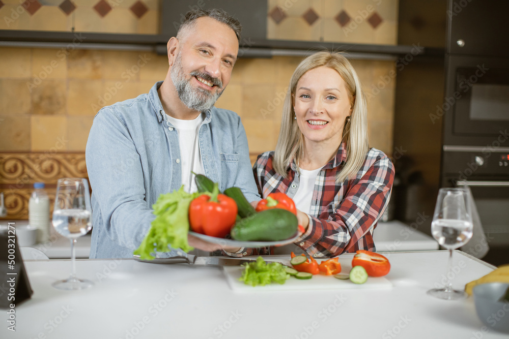 Portrait of beautiful mature couple standing on bright kitchen and holding plate with fresh organic vegetables. Healthy eating and lifestyles concept. Portrait of beautiful mature couple standing on bright kitchen and holding plate with fresh organic vegetables. Healthy eating and lifestyles concept.