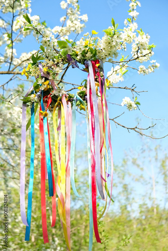flower wreath with colorful ribbons on tree in Spring garden, natural background. floral decor, Symbol of Beltane, Wiccan Celtic Holiday beginning of summer. pagan witch traditions, rituals