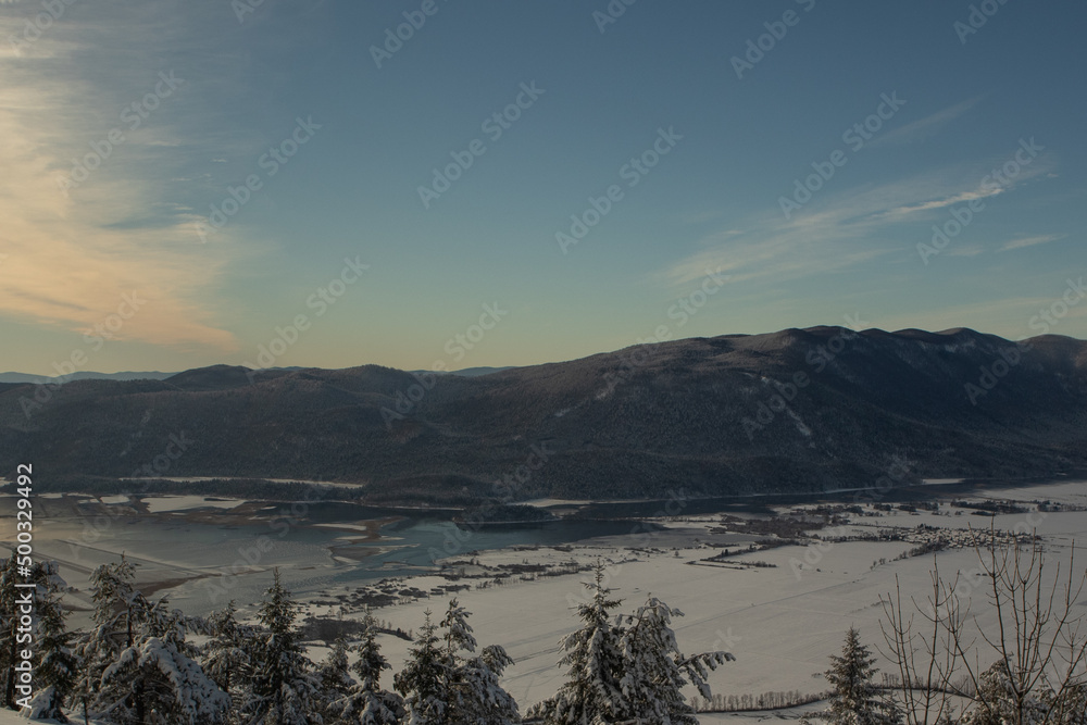 View from slivnica hill towards the Cerknica valley and Kalic ridge in winter time. Snow covered mountains and hills below, trees in foreground.
