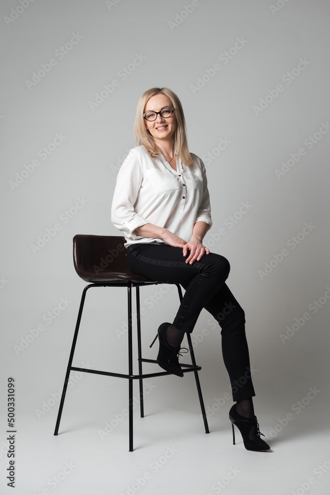 Smiling beautiful young woman with glasses sitting on a chair in a studio with grey background. Sales agent, manager, agent, assistant, teacher, student Smiling beautiful young woman with glasses sitting on a chair in a studio with grey background. Sales agent, manager, agent, assistant, teacher, student