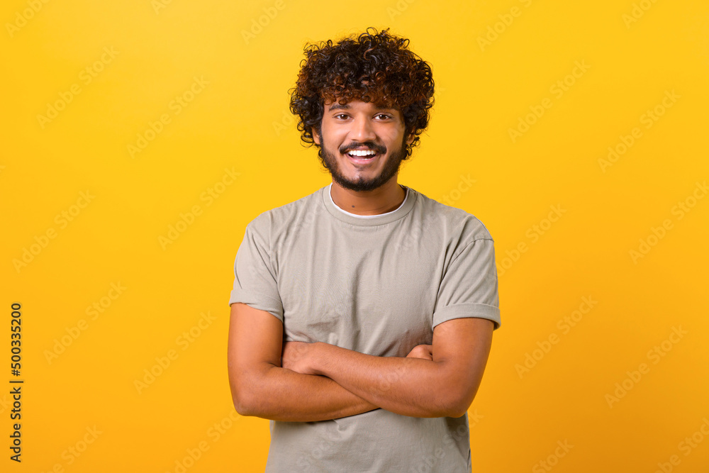 Indian youngster looking at camera standing with arms crossed isolated on yellow background. Eastern male student with curly hair in casual t-shirt with folded hands Indian youngster looking at camera standing with arms crossed isolated on yellow background. Eastern male student with curly hair in casual t-shirt with folded hands