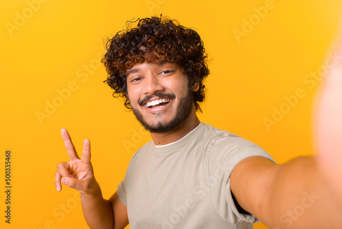 Portrait of young Indian handsome man taking selfie, looking at camera POV and gesturing hand, curly guy recording himself. Indoor studio shot isolated on white background