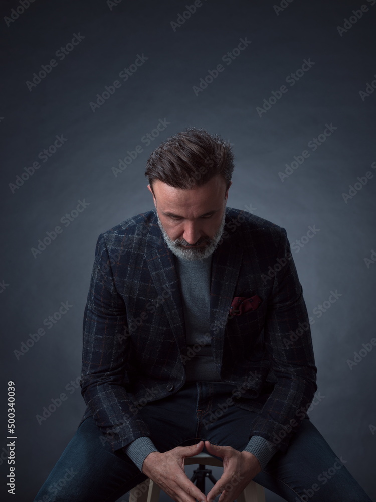 Portrait of adult businessman wearing trendy suit and sitting in modern studio on stylish chair against the black background