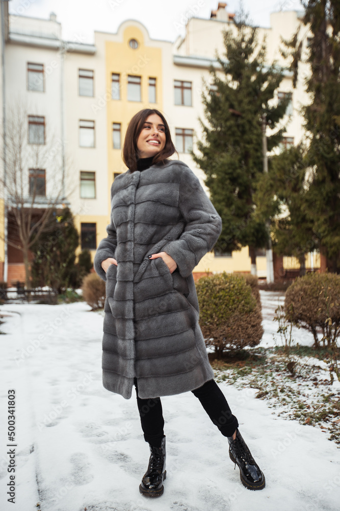 Girl posing on road on winter background. Glamorous funny young woman with smile wearing stylish grey long fur coat . Fur and fashion concept.