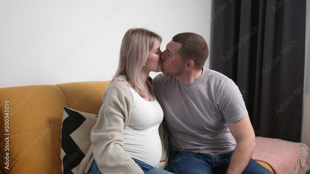 A couple expecting a baby are hugging at home on the couch. A man touches the belly of a pregnant wife sitting on the sofa in the living room. Young couple kissing A couple expecting a baby are hugging at home on the couch. A man touches the belly of a pregnant wife sitting on the sofa in the living room. Young couple kissing