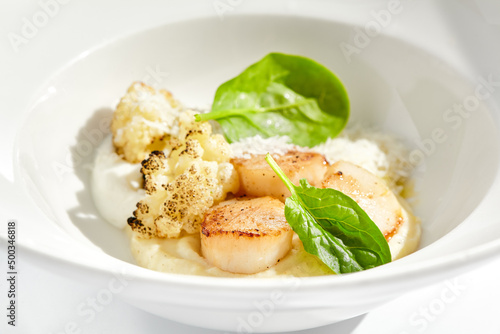 Gourmet dish - pan seared sea scallops with mashed potatoes, grilled cauliflowers on white plate. Scallops with vegetables and creamy espuma. Delicacy seafood in restaurant menu.
