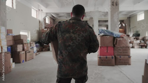Military man carries a heavy cardboard package and puts it on the pile of boxes in the humanitarian war centre.