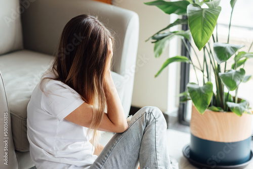 Unhappy young caucasian woman with blonde hair thinking about bad relationships problems, break up with boyfriend. Worried millennial girl sitting on floor in bedroom near chair and green plant alone photo
