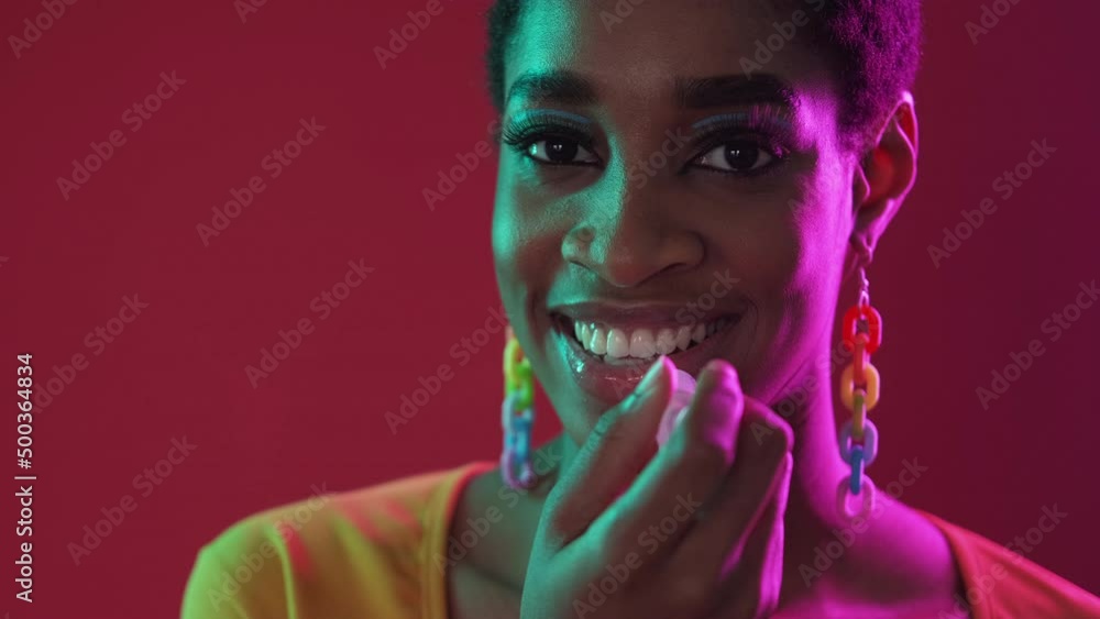 Party makeup. Lip moisturizing. Skincare cosmetic product. Confident smiling woman with perfect face skin applying gloss in color neon light isolated on dark red copy space background. Party makeup. Lip moisturizing. Skincare cosmetic product. Confident smiling woman with perfect face skin applying gloss in color neon light isolated on dark red copy space background.