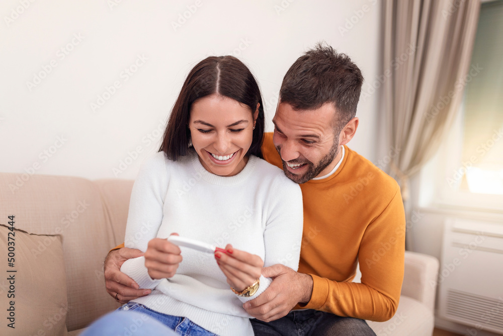 Enamoured couple finding out results of a pregnancy test sitting on the bed. Happy couple checking pregnancy test sitting on a couch in the living room at home. Family, parenting and medical concept Enamoured couple finding out results of a pregnancy test sitting on the bed. Happy couple checking pregnancy test sitting on a couch in the living room at home. Family, parenting and medical concept