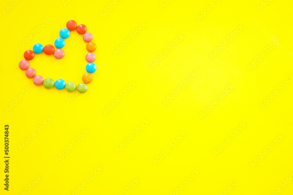 Sweet multi-colored chocolates, laid out in the shape of a heart. Yellow background. Space for text. Sweet multi-colored chocolates, laid out in the shape of a heart. Yellow background. Space for text.