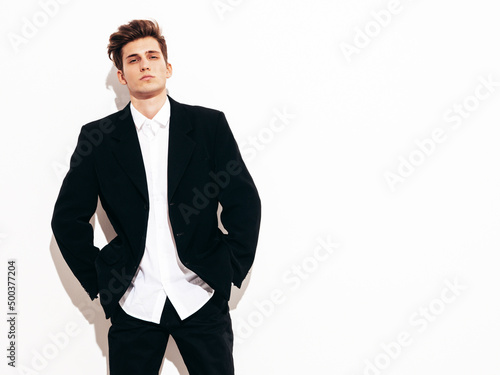 Portrait of handsome confident stylish hipster lambersexual model. Sexy modern man dressed in elegant black suit. Fashion male posing in studio near white wall. Isolated