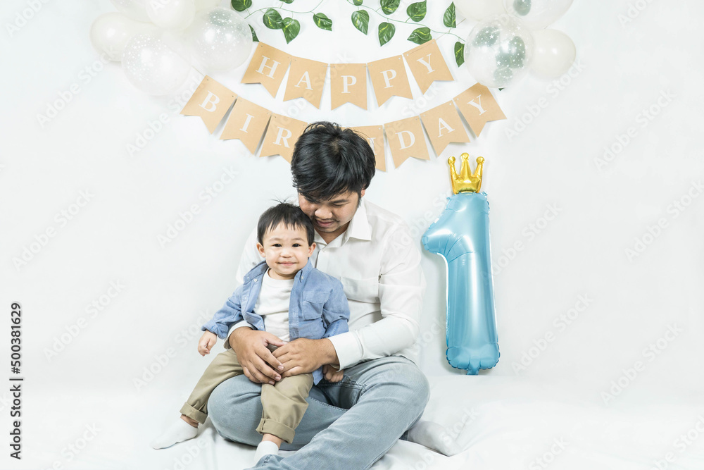 Naklejka premium Asian baby boy celebrating first birthday,A father sits and hug his son smiling on a floor with a minimal background,white balloons and birthday flag.Selective focus