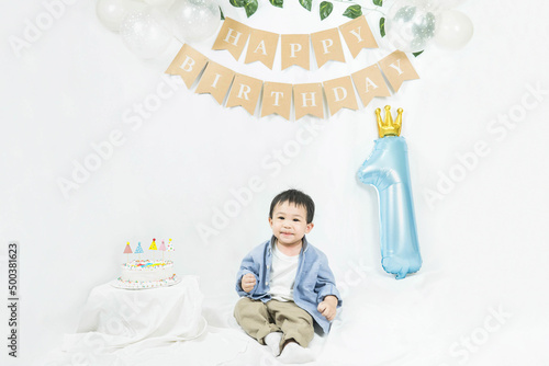 Asian baby boy celebrating first birthday,Cake for 1 year.Infant, small cute child dressed in t-shirt and blue shirt sitting on the floor with a minimal background,white balloons and birthday flag.