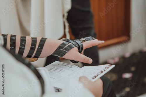 Jewish Man wrapped in tefillin prays. A religious orthodox Jew with arm-tefillin on his left hand, prays. Jewish teenager 13 years old celebrates bar mitzvah reads Jewish bible book