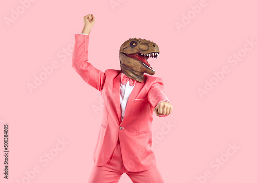 Funny confident man wearing rubber dinosaur mask dancing at show or event. Happy crazy T Rex guy in funky pink party suit with bow tie doing gangnam style moves isolated on pink colour background