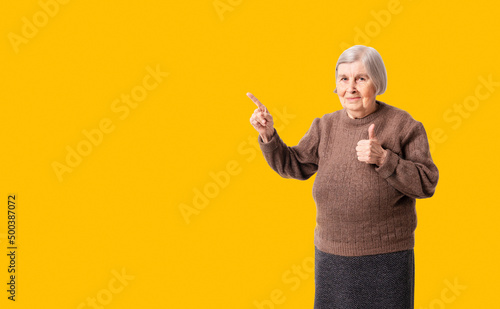 Cheerful octogenarian woman wearing brown sweater showing on empty place for copy space. Background for your promo or advertisement. Studio shoot.