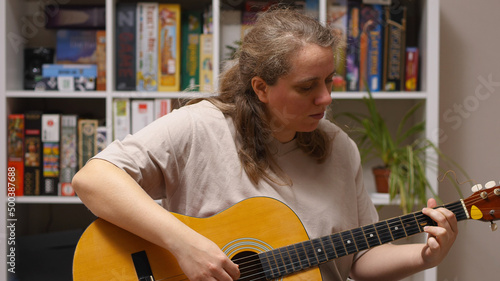 A girl plays an overdub on an acoustic guitar sitting in a room. Behind her is a rack of board games and flowers. A mash-up of music. photo