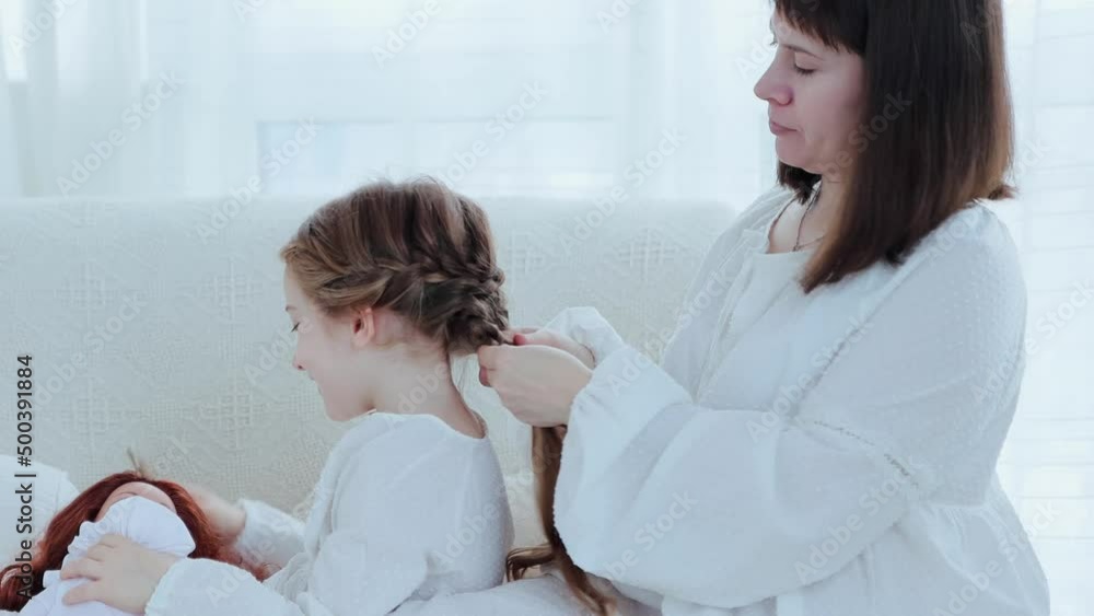 Young happy mother and little cheerful daughter sits on the couch while mom braids child's hair, side view 4k video Young happy mother and little cheerful daughter sits on the couch while mom braids child's hair, side view 4k video