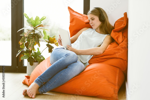 Photos barefoot caucasian girl sitting on bean bag chair with tablet and earphones at h