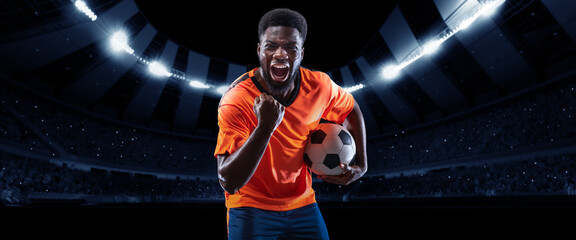 Victory. Excited professional soccer, football player in football kit standing with ball and shouting over dark night stadium with flashlights background.