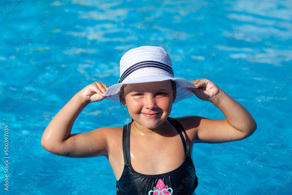 Child in white panama hat looks at camera. Happy charming tanned girl in black swimsuit stand in water and holds white straw hat decorated with ribbon in her hands and enjoys swimming lesson in pool. 