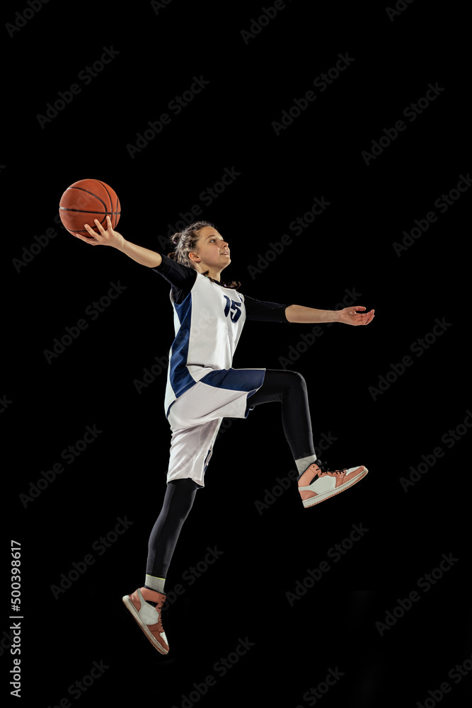 Jump shot. Scoring a goal. Portrait of teen girl, basketball player in motion, training, playing isolated over black background.