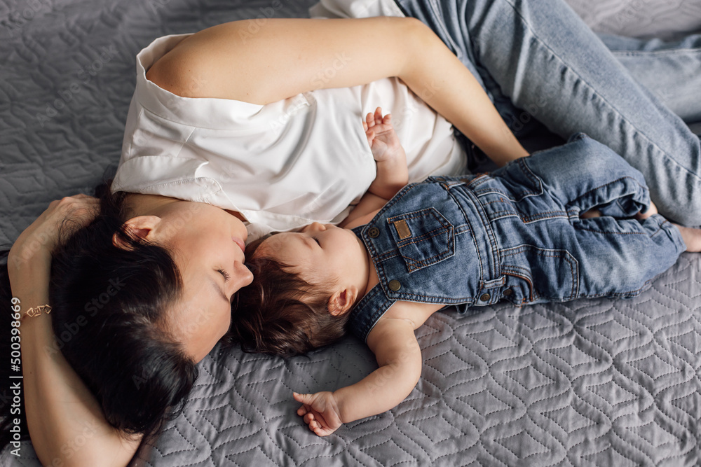 Portrait of young mother with little daughter in bedroom closeup, free copy space, gray background. Cozy lay on bed with baby, capture happy emotions. Concept of maternal affection and childcare.