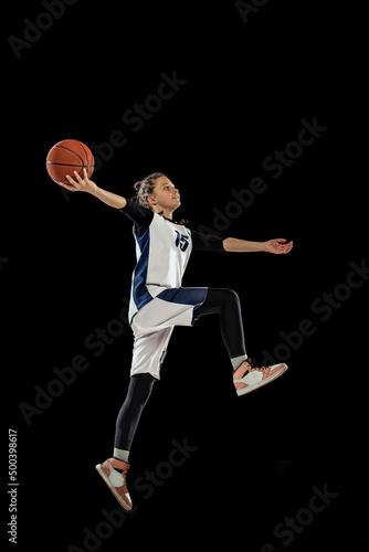 Jump shot. Scoring a goal. Portrait of teen girl, basketball player in motion, training, playing isolated over black background.