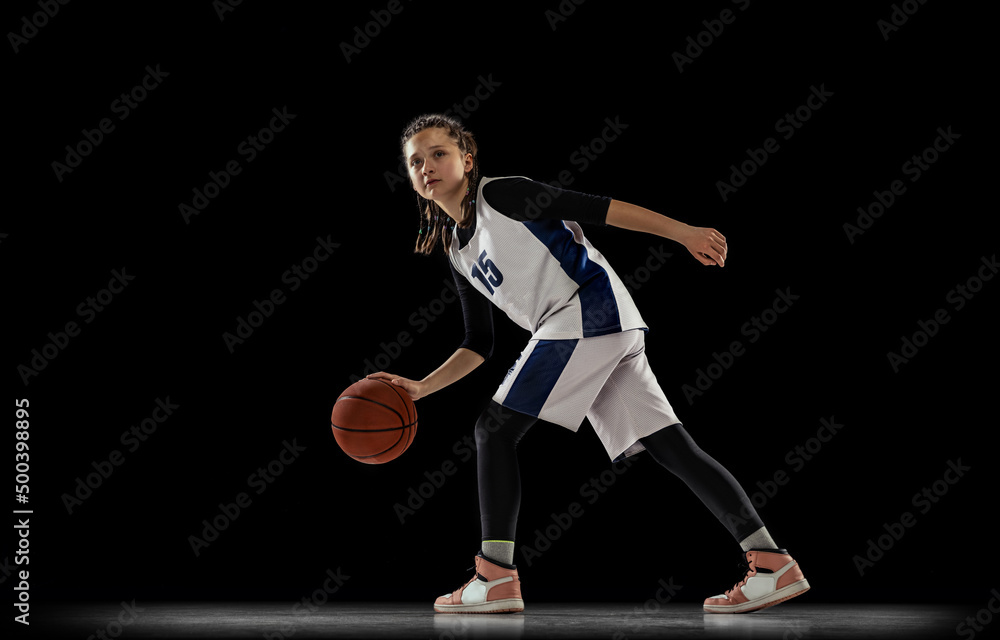 Portrait of young girl, basketball player in motion, training isolated over black background. Dribbling exercises.