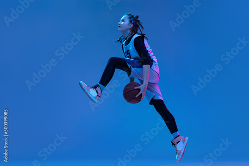 Dynamic portrait of teen girl, basketball player training, jumping with ball isolated over blue studio background in neon. Defender © Lustre
