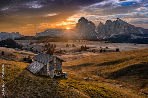 Alpe di Siusi, Italy - Aerial panoramic view of a wooden cottage at Seiser Alm, a Dolomite plateau in South Tyrol province in the Dolomites mountain range at autumn sunrise with colorful golden sky © zgphotography