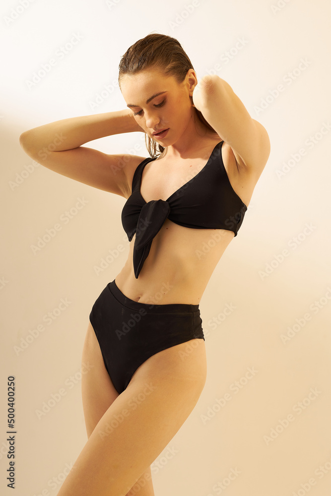 Obraz premium Fashion photo of young lady with perfect body in a black designer trend 2022 swimsuit. Girl on a sunny day sunbathes in sexy pose on the white. Sun screen, summer concept.