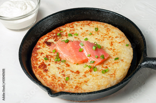 traditional Swiss dish is a potato pancake in a frying pan with pieces of red salmon fish, decorated with green onions. there is a gravy boat with white cream sauce nearby. close-up.