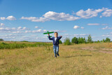 A six-year-old preschooler boy in a blue jacket launches a toy plane in a field against a blue sky with clouds on a summer day. The bright sun is shining. Scenery