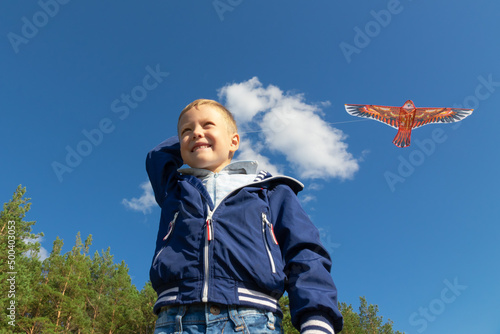 A six-year-old preschooler boy in a blue jacket launches a kite bird in nature against the background of a clear blue sky on a summer day. The bright sun is shining. Portrait