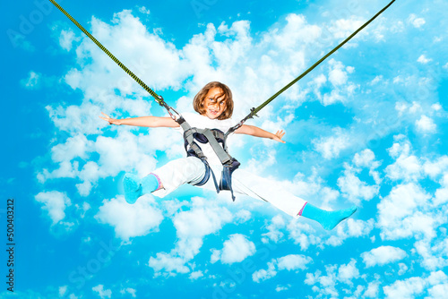 Little girl bouncing high in the air using a bungee trampoline against blue sky with white clouds.