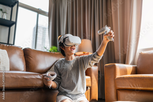 exiting asian child male boy enjoy metaverse gaming with wearable vr headset with control handle playing sport gaming online in living room at home,home technology young teen using vr technology