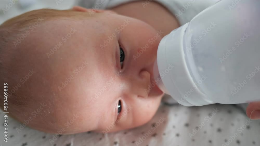 Newborn baby girl with half closed eyes drinks water from plastic bottle lying on bed. Mother feeds cute little kid at home closeup, side view Newborn baby girl with half closed eyes drinks water from plastic bottle lying on bed. Mother feeds cute little kid at home closeup, side view