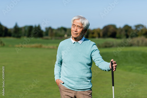 asian senior man standing with hand in pocket and holding golf club on green field.