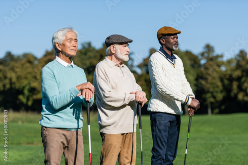 senior man in flat cap near interracial friends standing with golf clubs. © LIGHTFIELD STUDIOS