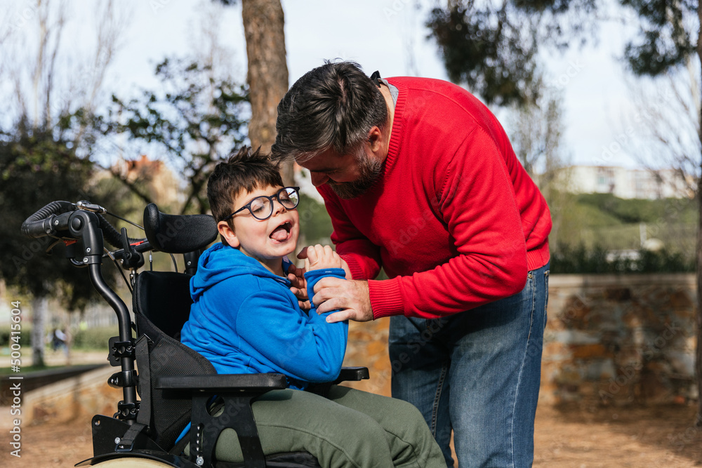 A father playing with his son with a disability who uses a wheelchair in the park A father playing with his son with a disability who uses a wheelchair in the park