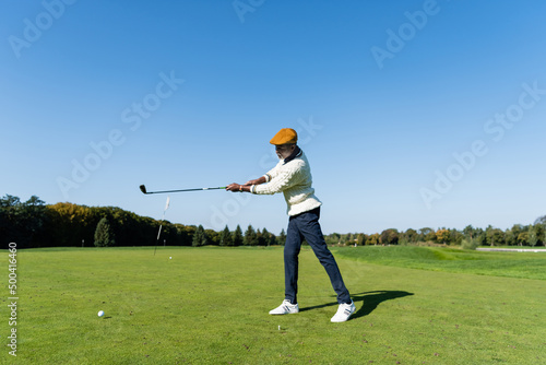 full length of african american man in flat cap holding golf club and playing on green lawn.