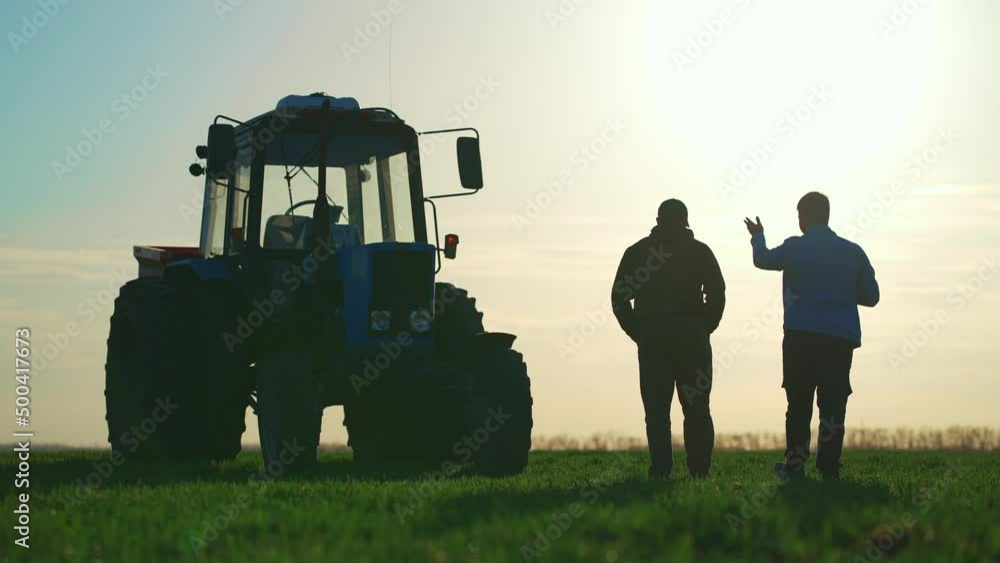Teamwork concept. Silhouette two male farmers walking in a green field against sunset. Team farmers stand in a field on the background of agricultural machinery. Agronomists discuss harvest.