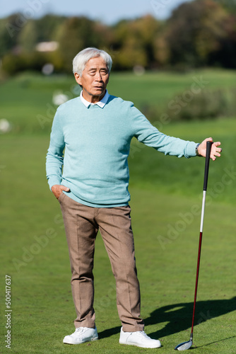 asian senior man standing with hand in pocket and golf club on green field.