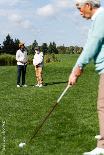 senior interracial men in flat caps looking at asian friend playing golf on blurred foreground.