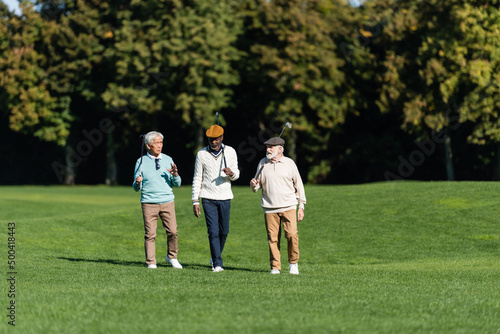 interracial senior friends walking with golf clubs on field.