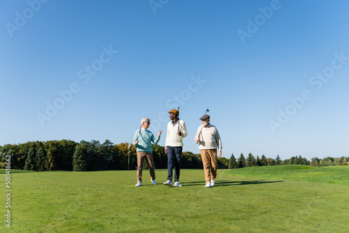 senior interracial friends walking with golf clubs on field.