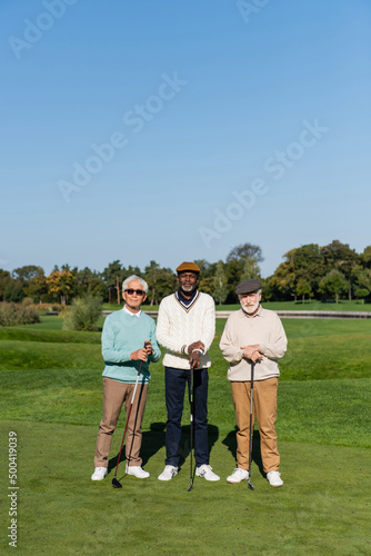 asian man in sunglasses standing near senior multiethnic friends in flat caps with golf clubs.